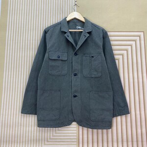 Könnte beinhalten: Dunkelgrüne Jacke mit vier Vordertaschen und Knopfverschluss. Die Jacke hat lange Ärmel und einen Kragen. Der Markenname "Gap" ist auf dem Etikett sichtbar. Die Jacke hängt an einem Holzbügel.