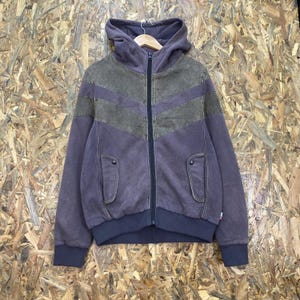 May include: A full-zip hooded jacket in muted purple and olive green. The garment features a chevron-patterned olive green panel across the chest, a central black zipper, two front pockets with snap closures, and ribbed cuffs and hem.