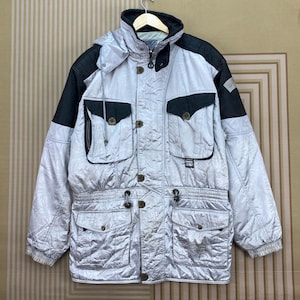 May include: A silver and black hooded winter jacket with multiple pockets and button closures. The jacket has a quilted pattern and a high collar. The sleeves and shoulders are black, and the jacket is hanging on a hanger.