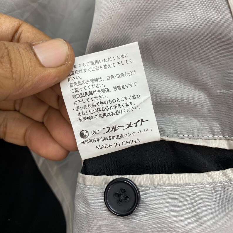 Puede incluir: Primer plano de una etiqueta de prenda con texto japon&eacute;s, un bot&oacute;n negro y un forro gris. La etiqueta dice "MADE IN CHINA" e incluye texto japon&eacute;s. El bot&oacute;n est&aacute; centrado en la parte inferior de la imagen.