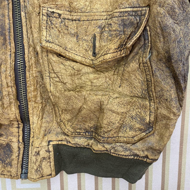 May include: Close-up of a distressed, tan leather jacket with a dark zipper and a pocket. The jacket has a worn, textured appearance with dark brown accents and a green ribbed hem.