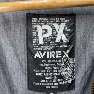 May include: Close-up of a black label with white text and the logo "AVIREX". The label includes text about garment care and specifications. The background is a gray fabric.