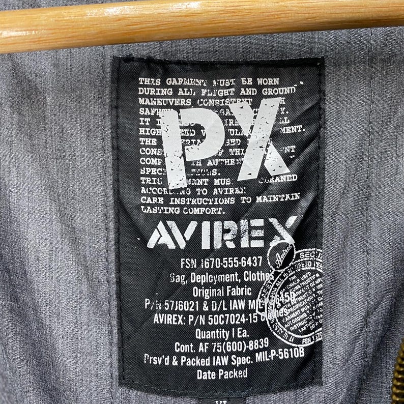 May include: Close-up of a black label with white text and the logo "AVIREX". The label includes text about garment care and specifications. The background is a gray fabric.