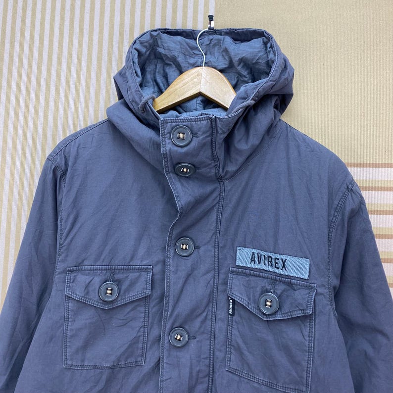 May include: A blue-gray hooded jacket with a button-up front and two front pockets. The jacket features the brand name "AVIREX" on a patch. The hood is attached and the jacket has a casual, utilitarian style.