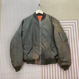 May include: Olive green bomber jacket with a contrasting orange lining. Features a zip-up front, two front pockets, and a zippered pocket on the left sleeve. The jacket has ribbed cuffs and a ribbed waistband. The jacket is hanging on a wooden hanger.