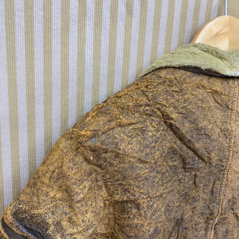 May include: Close-up of a vintage brown leather jacket with a distressed texture. The jacket features a light green faux fur collar and is hanging on a wooden hanger. The leather has a worn, aged appearance.