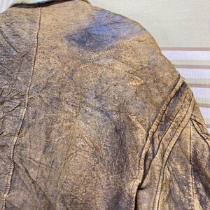 May include: Close-up of a vintage brown leather jacket. The jacket has a distressed, textured appearance with visible wear and tear. The collar is visible, and the jacket appears to be well-worn, with a classic design.