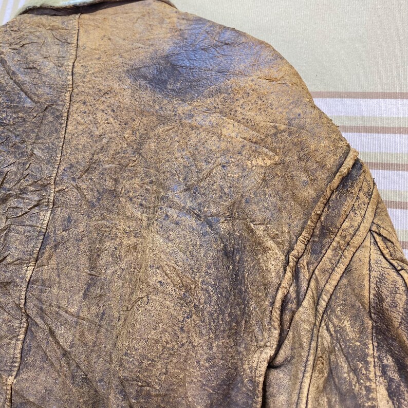 May include: Close-up of a vintage brown leather jacket. The jacket has a distressed, textured appearance with visible wear and tear. The collar is visible, and the jacket appears to be well-worn, with a classic design.