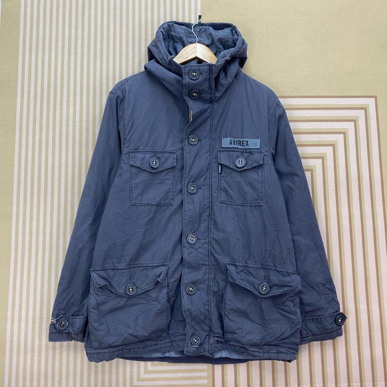 May include: A blue hooded jacket with multiple front pockets and button closures. The jacket features the "AVIREX" logo on the chest and a buttoned cuff detail. The jacket is made of a durable material.