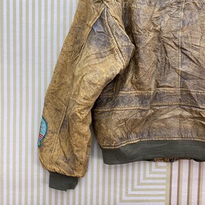 May include: A vintage brown leather bomber jacket with a distressed finish. The jacket features a green ribbed hem and cuffs, and a small patch on the left sleeve. The leather has a worn, textured appearance.