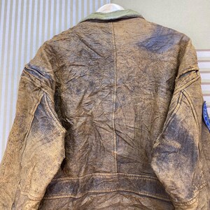 May include: A vintage brown leather jacket with a distressed appearance. The jacket features a green collar and a blue patch on the sleeve. The leather shows signs of wear, adding to its classic, rugged aesthetic.
