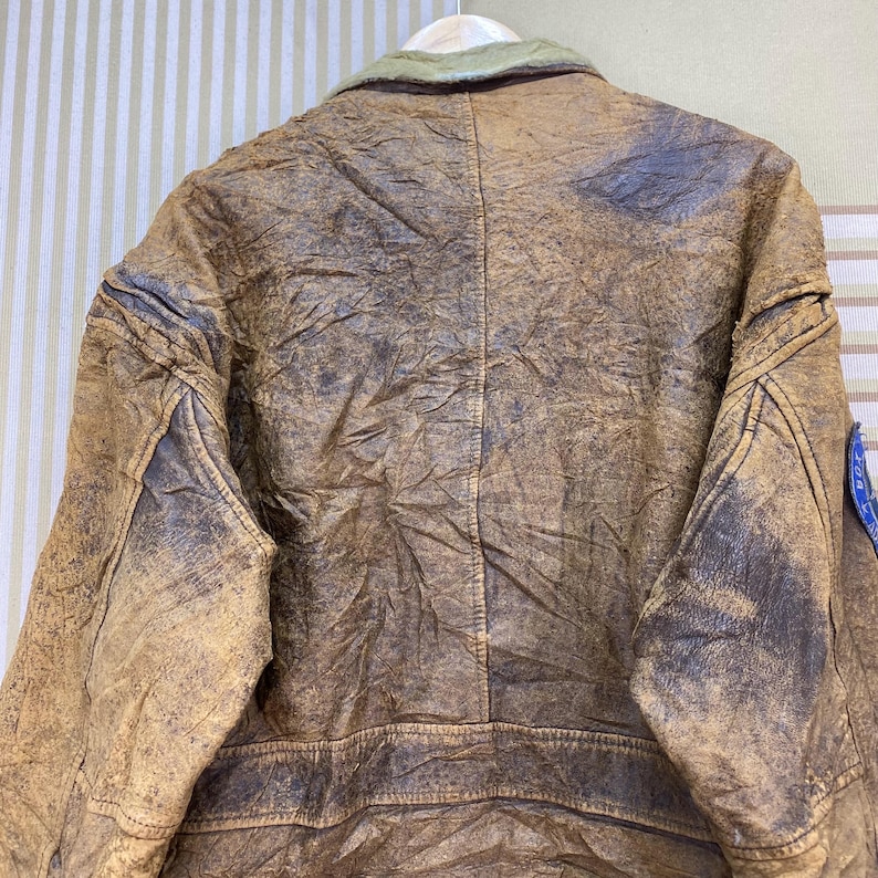 May include: A vintage brown leather jacket with a distressed appearance. The jacket features a green collar and a blue patch on the sleeve. The leather shows signs of wear, adding to its classic, rugged aesthetic.