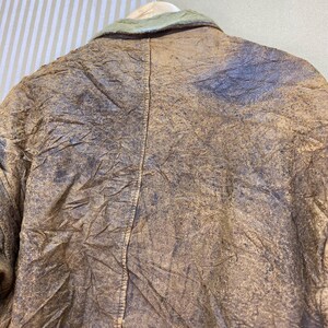 May include: Close-up of a vintage brown leather jacket with a light green collar. The jacket has a distressed appearance with visible creases and variations in color, suggesting age and wear. The jacket is hanging on a wooden hanger.