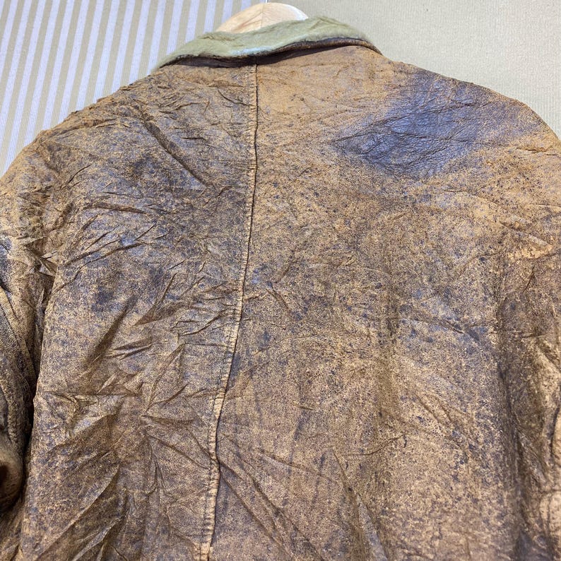 May include: Close-up of a vintage brown leather jacket with a light green collar. The jacket has a distressed appearance with visible creases and variations in color, suggesting age and wear. The jacket is hanging on a wooden hanger.