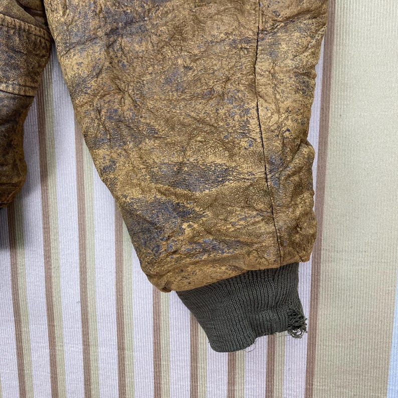 May include: Close-up of a vintage brown leather jacket sleeve. The leather has a distressed, textured appearance with dark gray accents. The sleeve ends with a worn, olive green ribbed cuff.