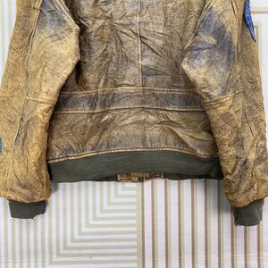 May include: A vintage brown leather jacket with a distressed appearance. The jacket features a green ribbed hem and cuffs, and patches on the sleeves. The leather has a worn, textured look.