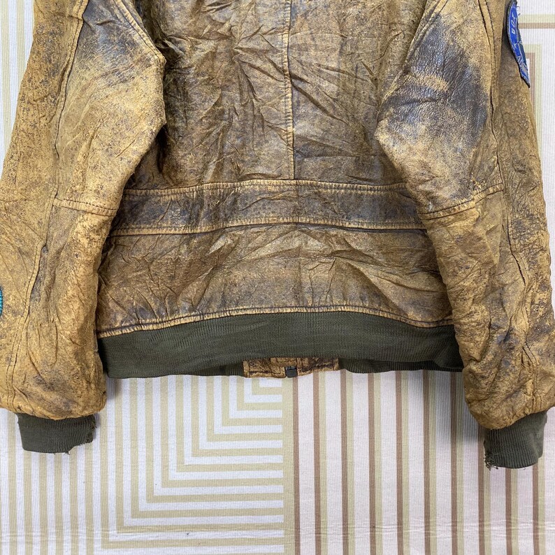 May include: A vintage brown leather jacket with a distressed appearance. The jacket features a green ribbed hem and cuffs, and patches on the sleeves. The leather has a worn, textured look.