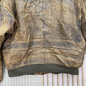 May include: Close-up of a vintage brown leather jacket with a distressed, textured appearance. The jacket features a ribbed olive green hem and cuffs. The leather shows signs of wear, with darker areas and visible stitching.
