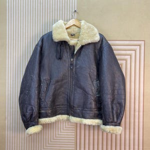 以下が含まれることがあります： 厚手のクリーム色のシープスキン裏地、幅広の折り返し襟、中央のフロントジッパー、2つのサイドポケットを備えたダークブラウンのレザーボンバージャケット。袖口と裾にはシープスキンのトリミングが施されています。