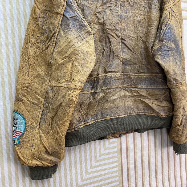 May include: A distressed brown leather jacket with a faded appearance. The jacket features a ribbed green hem and cuffs, and a patch on the left sleeve with a design in blue, red, and white. The jacket has a vintage, worn look.