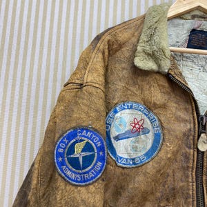 May include: A vintage brown leather jacket with two embroidered patches. One patch reads "Box Canyon Administration" and the other says "USS Enterprise Van 65". The jacket has a shearling collar and a map-like lining.
