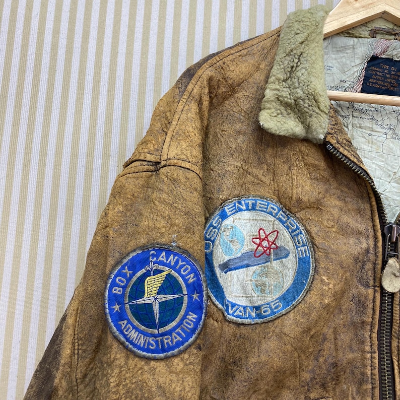 May include: A vintage brown leather jacket with two embroidered patches. One patch reads "Box Canyon Administration" and the other says "USS Enterprise Van 65". The jacket has a shearling collar and a map-like lining.