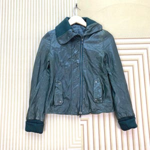 Pode incluir: Casaco de couro verde escuro com gola alta e fecho de correr. O casaco tem dois bolsos frontais com botões e punhos verde escuro. Está pendurado num cabide de madeira.