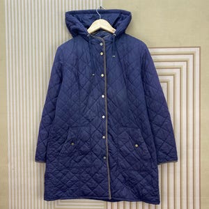 Puede incluir: Una chaqueta acolchada de color azul marino con capucha, cierre frontal de botones a presión plateados y dos grandes bolsillos de parche. La chaqueta tiene un fino borde marrón a lo largo de la abertura frontal y cuelga de una percha.