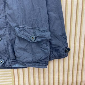 May include: Close-up of a blue jacket with two front pockets and button closures. The jacket has a textured appearance and a buttoned cuff detail. The fabric appears to be a lightweight material, suitable for layering.