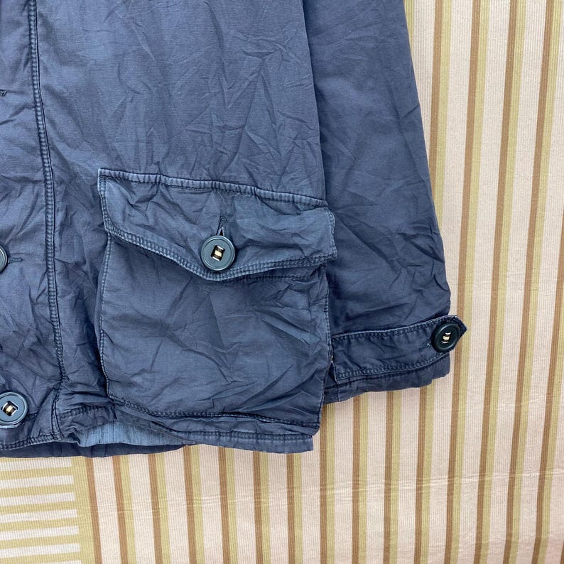 May include: Close-up of a blue jacket with two front pockets and button closures. The jacket has a textured appearance and a buttoned cuff detail. The fabric appears to be a lightweight material, suitable for layering.