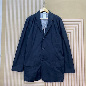 Puede incluir: Un blazer negro con solapa de muesca, tres botones y dos bolsillos delanteros. El blazer est&aacute; colgado de una percha de madera sobre un fondo a rayas. El blazer parece estar hecho de una tela ligera.
