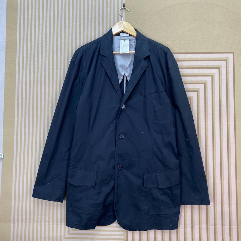 Puede incluir: Un blazer negro con solapa de muesca, tres botones y dos bolsillos delanteros. El blazer est&aacute; colgado de una percha de madera sobre un fondo a rayas. El blazer parece estar hecho de una tela ligera.