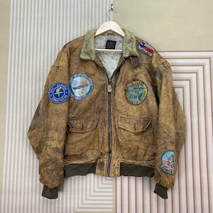 May include: Brown leather bomber jacket with multiple patches, including "Dallas" and "Tomca." The jacket has a fur collar, two front pockets, and a green ribbed hem and cuffs. The jacket is hanging on a wooden hanger.