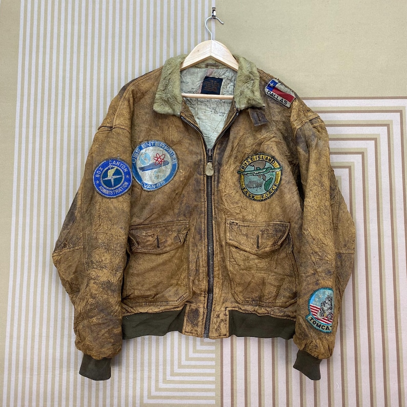 May include: Brown leather bomber jacket with multiple patches, including "Dallas" and "Tomca." The jacket has a fur collar, two front pockets, and a green ribbed hem and cuffs. The jacket is hanging on a wooden hanger.