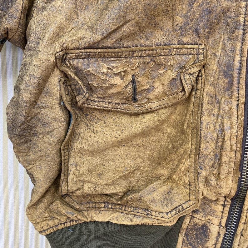May include: Close-up of a distressed, tan leather jacket with a large, square pocket. The jacket has a worn, textured appearance with visible stitching and a dark green ribbed cuff. A metal zipper is visible on the right side.