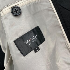Puede incluir: Primer plano del forro interior de una chaqueta negra. El forro es de color gris claro con un bot&oacute;n negro y una etiqueta que dice "SACSNY Y SACCS" con una talla "4L".