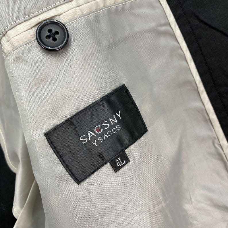 Puede incluir: Primer plano del forro interior de una chaqueta negra. El forro es de color gris claro con un bot&oacute;n negro y una etiqueta que dice "SACSNY Y SACCS" con una talla "4L".