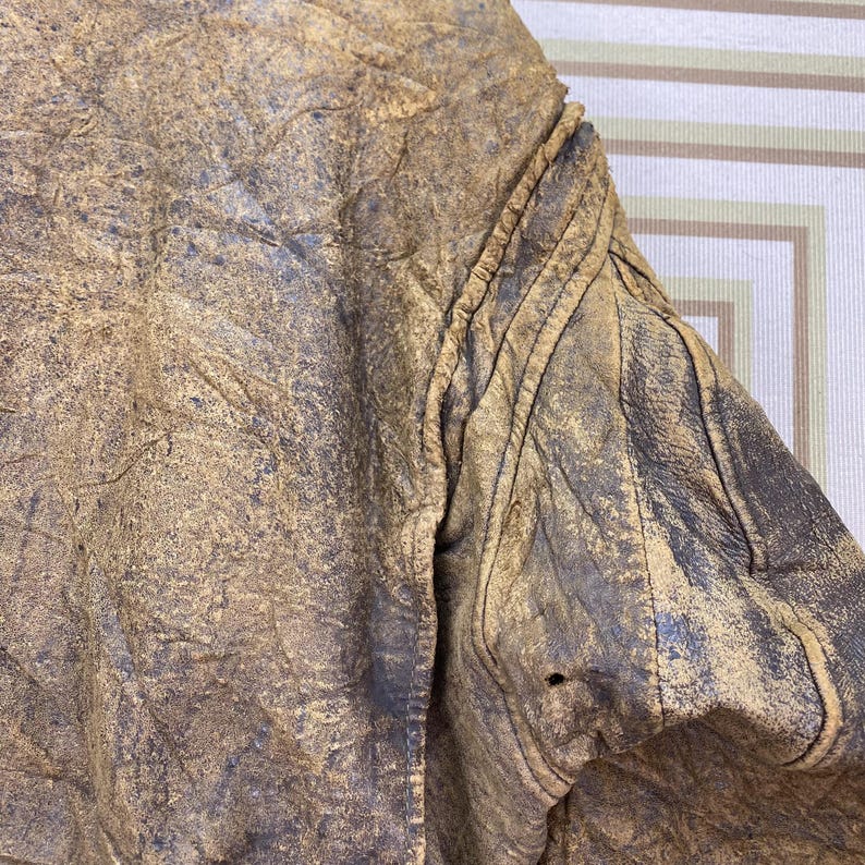 May include: Close-up of a distressed brown leather jacket. The jacket has a worn, textured appearance with visible creases and a small hole. The shoulder area features stitched detailing.