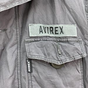 May include: Close-up of a gray jacket with a pocket and the brand name "AVIREX" visible on a patch and a label. The jacket has a button closure and visible stitching details.