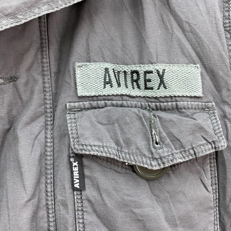 May include: Close-up of a gray jacket with a pocket and the brand name "AVIREX" visible on a patch and a label. The jacket has a button closure and visible stitching details.