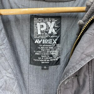 May include: Close-up of a gray jacket with a hood and a black label. The label features the text "PX" and "AVIREX", along with other text and numbers. The jacket is size XL.