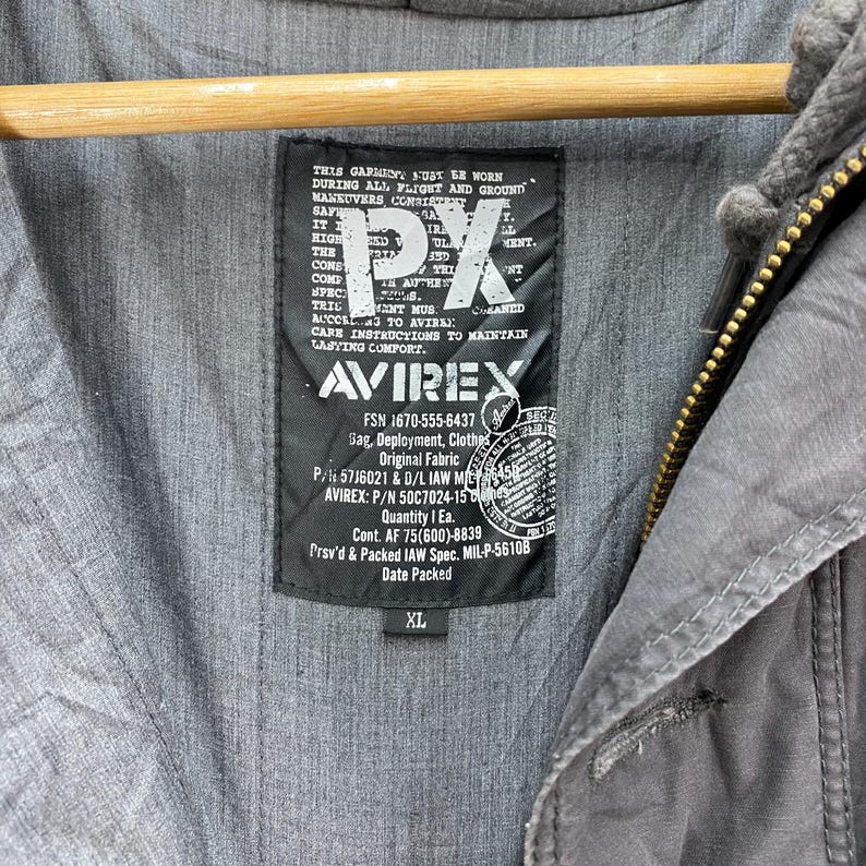 May include: Close-up of a gray jacket with a hood and a black label. The label features the text "PX" and "AVIREX", along with other text and numbers. The jacket is size XL.
