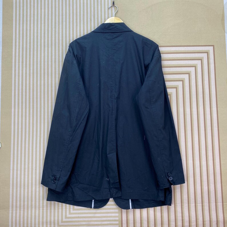 Puede incluir: Un blazer negro colgado de una percha de madera. El blazer tiene un dise&ntilde;o cl&aacute;sico con solapa de muesca, mangas largas y pu&ntilde;os abotonados. La parte trasera del blazer es visible, mostrando un corte entallado y una abertura en la parte inferior. El blazer es de tela oscura.