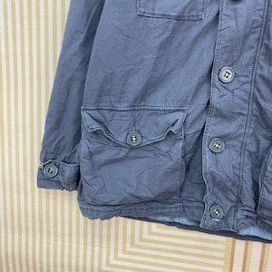 May include: A dusty blue jacket with multiple pockets and button closures. The jacket features a classic design with a collared neckline and buttoned cuffs. The buttons are a darker shade of blue, providing contrast. The fabric appears to be a durable material.