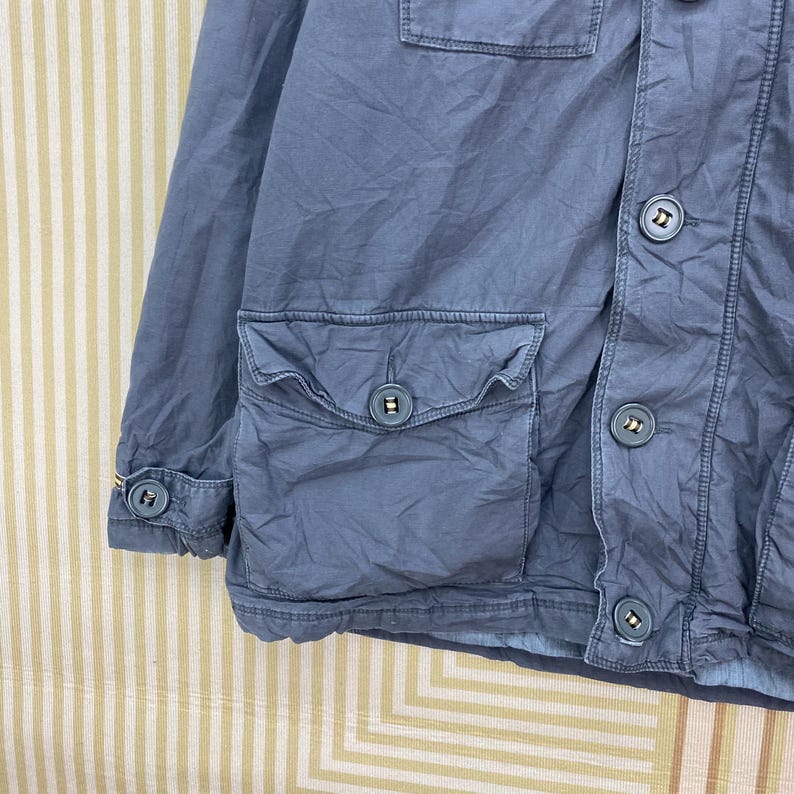 May include: A dusty blue jacket with multiple pockets and button closures. The jacket features a classic design with a collared neckline and buttoned cuffs. The buttons are a darker shade of blue, providing contrast. The fabric appears to be a durable material.