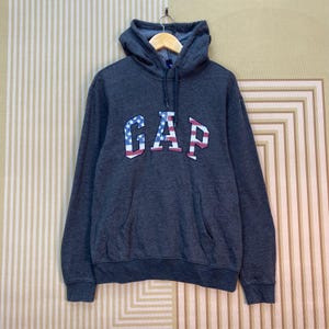 Puede incluir: Sudadera con capucha gris oscuro con la palabra "GAP" en un diseño patriótico rojo, blanco y azul en el pecho. La sudadera tiene un bolsillo de canguro y una capucha con cordón. La prenda está colgada en una percha.