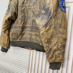 May include: A distressed brown leather jacket with a green ribbed hem and cuffs. The jacket has a worn appearance with visible stitching and patches. A circular patch with text is visible on the upper arm.