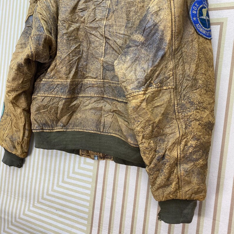 May include: A distressed brown leather jacket with a green ribbed hem and cuffs. The jacket has a worn appearance with visible stitching and patches. A circular patch with text is visible on the upper arm.