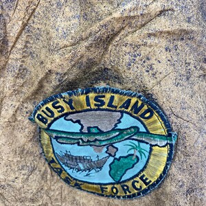May include: Embroidered patch with the text "BUSY ISLAND TASK FORCE" in gold lettering. The patch features a map, a ship, and a palm tree on a blue background, with a gold border. The patch is on a textured, brown surface.