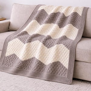 Puede incluir: Una manta con estampado de chevrón gris, crema y blanco sobre un sofá beige. La manta tiene un aspecto texturizado, tejido, con un borde gris liso. El sofá está en un salón con una alfombra y una mesa auxiliar.